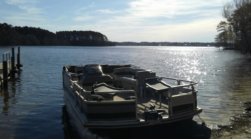 A pontoon boat docked by a serene lake, with sunlight reflecting on the water and trees in the background.