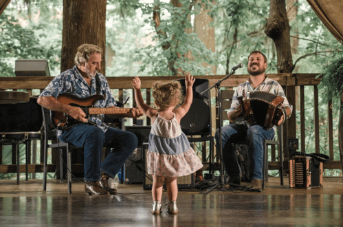 A young girl dances joyfully in front of two musicians playing guitar and accordion in a forest setting.
