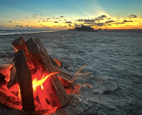 A glowing campfire on the beach at sunset, with a truck parked in the background and colorful clouds in the sky.
