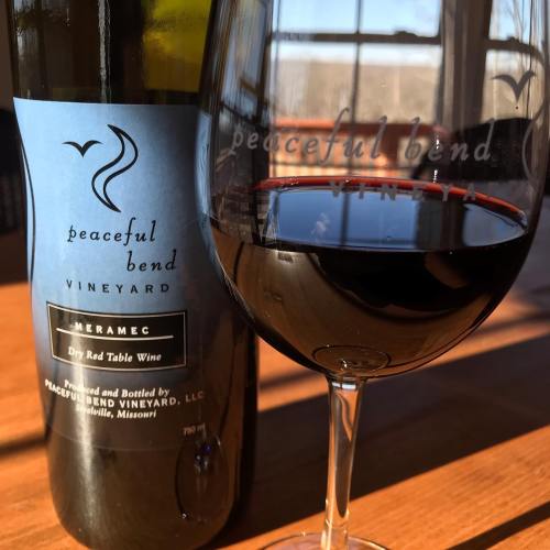 A glass of red wine beside a bottle labeled "Peaceful Bend Vineyard Merlot" on a wooden table.
