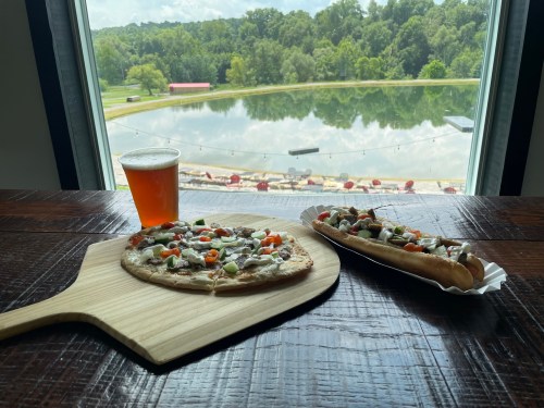 A wooden table with a pizza and hot dog, alongside a beer, overlooking a scenic lake and green landscape.
