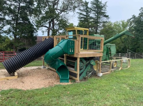 A green playground structure resembling a vintage tractor with slides and climbing features, set in a grassy area.
