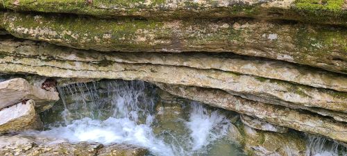 A small waterfall cascades over layered rock formations, surrounded by moss and greenery.