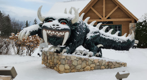 A large, snow-covered monster statue with sharp teeth and spikes, positioned outside a wooden building.