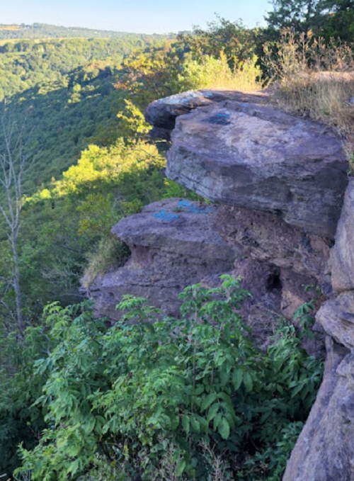 A rocky cliff edge overlooking a lush green valley and distant hills under a clear blue sky.