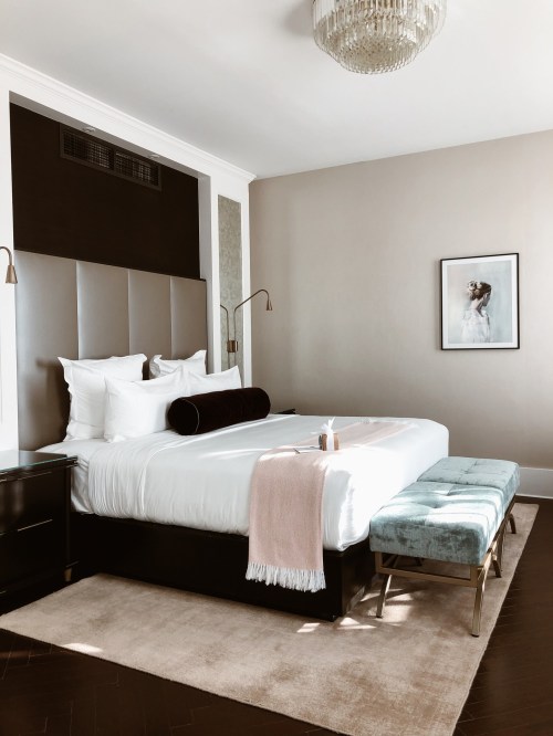 A stylish hotel room featuring a large bed, decorative pillows, a plush bench, and elegant lighting.