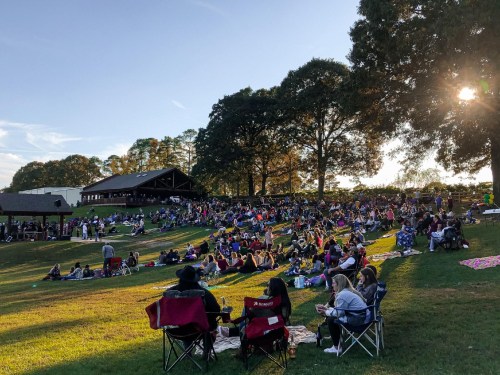 A large crowd enjoys an outdoor event on a grassy hill, with people sitting on blankets and chairs under trees.