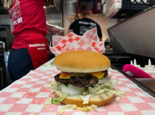 A close-up of a cheeseburger with lettuce, tomato, and onion on a counter, with staff preparing food in the background.