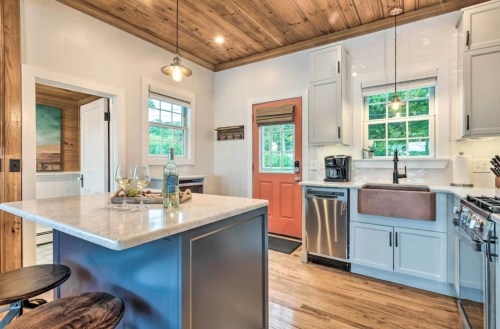 Bright kitchen with wooden ceiling, white cabinets, a farmhouse sink, and an orange door, featuring a marble island and bar stools.