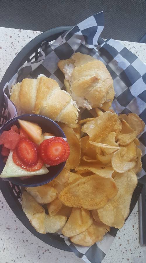 A basket with two croissant sandwiches, a side of potato chips, and a small bowl of mixed fruit.