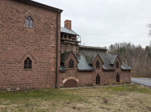 Historic stone building with gothic windows, featuring a sloped roof and a chimney, set against a cloudy sky.