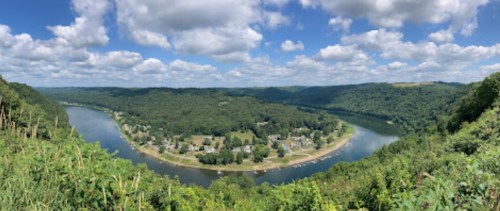 A panoramic view of a river winding through lush green hills under a blue sky with fluffy clouds.