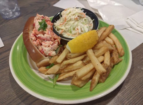A plate with a lobster roll, coleslaw, lemon wedge, and a side of French fries on a green dish.