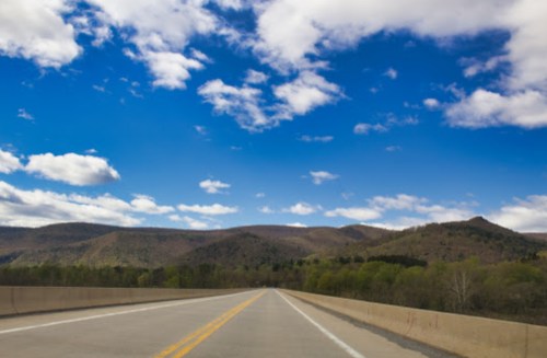 A wide road stretches into the distance, flanked by green hills under a blue sky with fluffy white clouds.