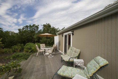 A patio with green chairs, a table, and an umbrella, surrounded by trees under a partly cloudy sky.
