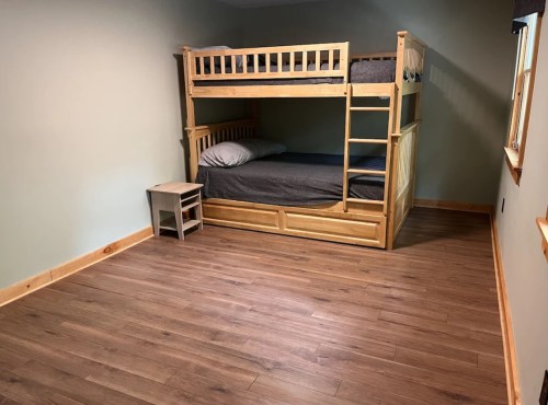 A simple bedroom featuring a wooden bunk bed and a small nightstand on a hardwood floor with green walls.