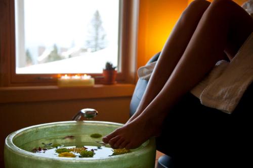 A person's feet resting in a green bowl filled with water and flowers, with a cozy room and window in the background.