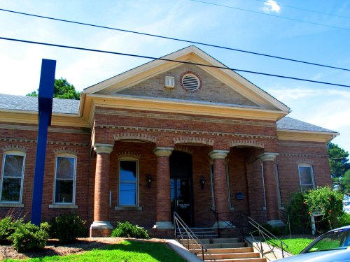 Historic brick building with a prominent entrance, columns, and a blue sign, surrounded by greenery and blue sky.