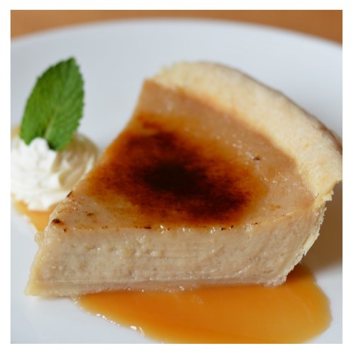 A slice of creamy dessert with a caramelized top, served with a dollop of whipped cream and a mint leaf on a white plate.