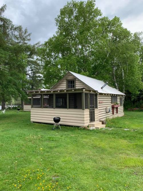 A small, beige cabin with a porch, surrounded by green grass and trees under a cloudy sky.