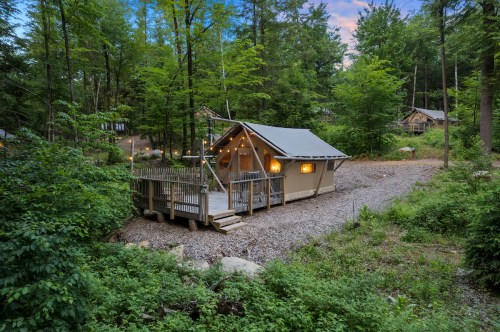 A cozy cabin nestled in a forest, surrounded by trees and gravel, with a deck and warm lights glowing from the windows.