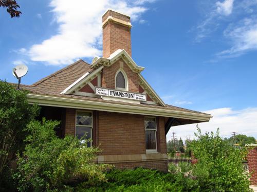 Historic brick train station with a chimney, sign reading "Evanston," surrounded by greenery and a blue sky.