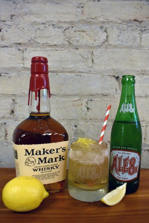 A bottle of Maker's Mark whiskey, a glass with a cocktail and straw, and an Ale-8 soda bottle, with a lemon on the side.