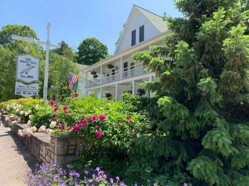 A charming inn surrounded by vibrant flowers and greenery, featuring a porch and an American flag. Established in 1896.