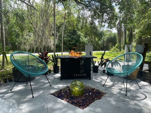 A cozy outdoor seating area with two teal chairs, a fire pit, and lush greenery in the background.