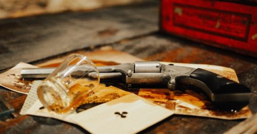 A revolver rests on vintage playing cards, next to a tipped glass with whiskey, on a wooden surface.