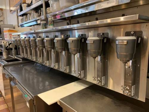 A row of coffee dispensers mounted on a stainless steel wall in a cafe kitchen.