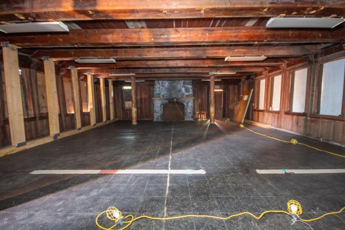Empty room with wooden beams, a stone fireplace, and black flooring, illuminated by yellow work lights.