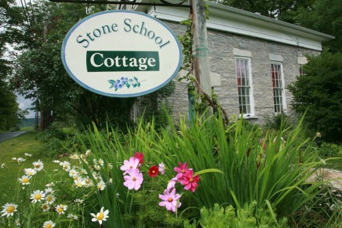 A sign reading "Stone School Cottage" in front of a stone building, surrounded by colorful flowers and greenery.