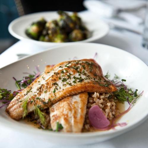 A plate of grilled fish on a bed of grains, garnished with herbs and pickled onions, with Brussels sprouts in the background.