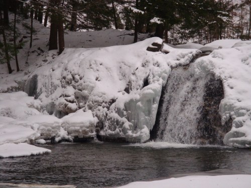 A frozen waterfall surrounded by snow-covered trees and icy terrain, with water flowing into a dark pool below.