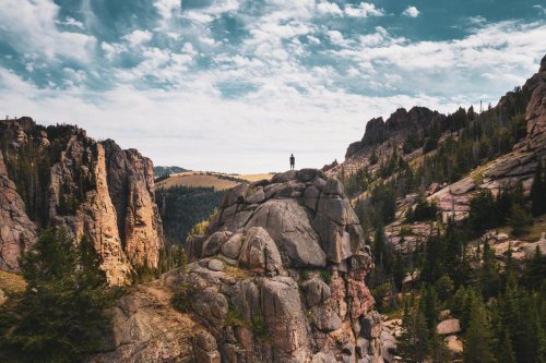 A person stands on a rocky outcrop, surrounded by towering cliffs and lush greenery under a blue sky with clouds.