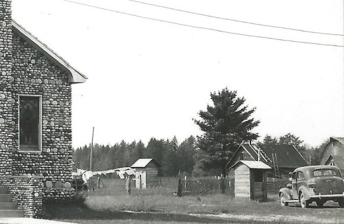 Black and white photo of a stone house, vintage car, and rustic buildings in a rural landscape.