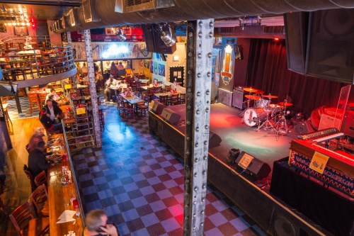 A lively music venue with a stage, seating areas, and a bar, featuring a vibrant atmosphere and checkered flooring.