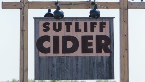 Sign for Sutliff Cider, featuring bold letters on a wooden background, hanging from a wooden post with lights above.