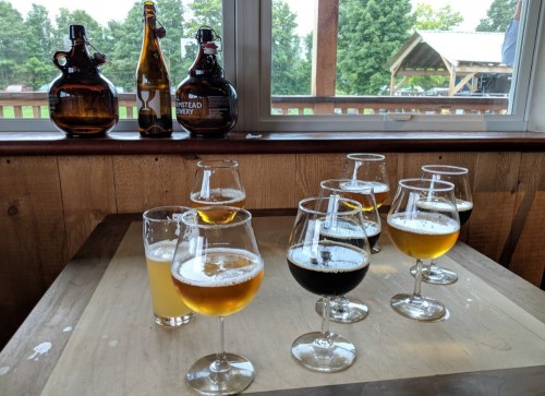 A table with various glasses of beer, including light and dark varieties, and two growlers in the background.