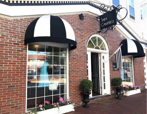 Exterior of a brick storefront with black and white awnings, featuring large windows and flower planters.