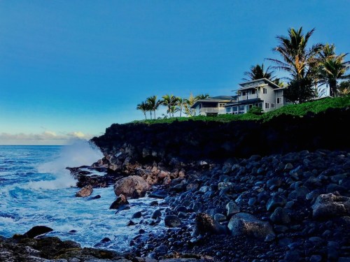 A coastal scene with a house on a cliff, palm trees, and waves crashing against rocky shore under a blue sky.