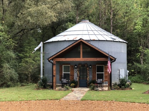 A unique round house with a metal roof, wooden porch, and American flag, surrounded by trees and a gravel path.