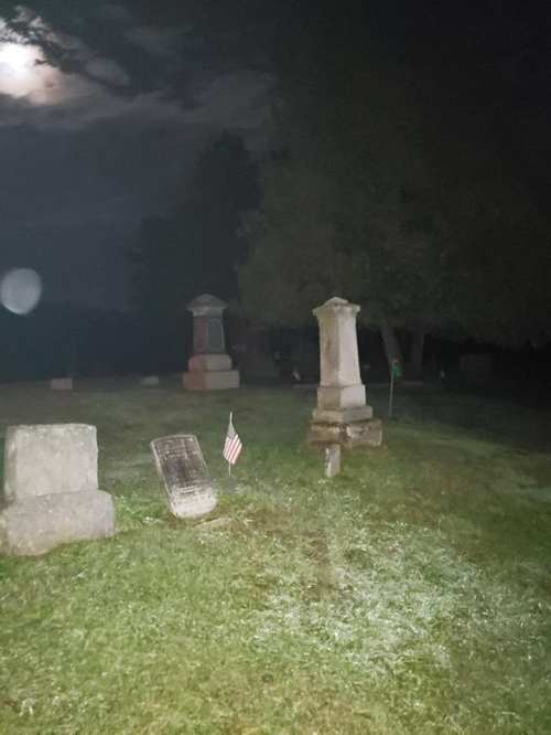A dimly lit cemetery at night, featuring gravestones and a small American flag on the grass.