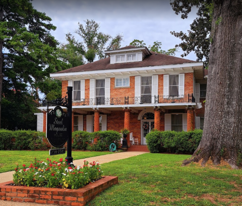 A large brick house with a porch, surrounded by greenery and flowers, featuring a sign for "Steel Magnolia."