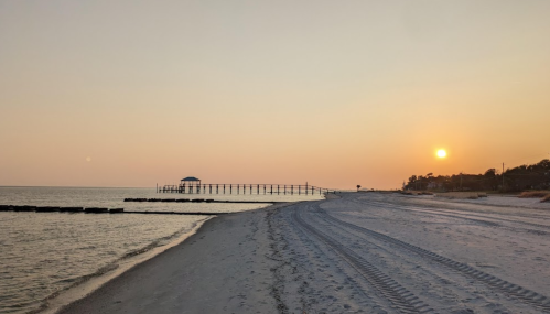 A serene beach at sunset, with gentle waves, a pier in the distance, and soft sand under a colorful sky.