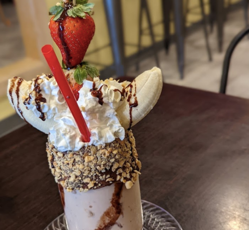 A decadent milkshake topped with whipped cream, a banana, a strawberry, chocolate drizzle, and crushed nuts.