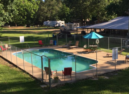 A fenced swimming pool surrounded by lounge chairs and an umbrella, with RVs and trees in the background.