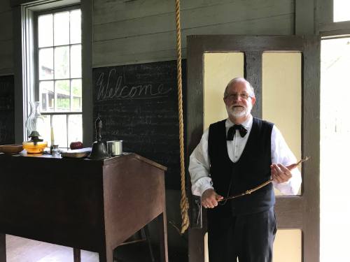 A man in period clothing stands in a historic room, holding a stick, with a welcome sign and chalkboard in the background.