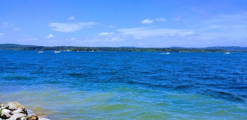 A serene blue lake under a clear sky, with distant boats and green hills in the background.
